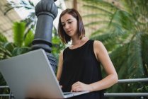 Low angle portrait of young brunette girl in black dress sitting near palm trees and using laptop on knees — Stock Photo