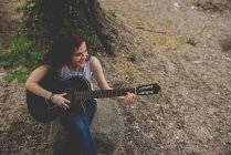 Retrato de ángulo alto de niña pecosa sonriente sentada en el rock y tocando la guitarra - foto de stock