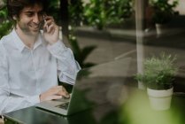 Portrait d'un homme d'affaires souriant assis à table et parlant sur smartphone tout en utilisant un ordinateur portable à la terrasse du café — Photo de stock