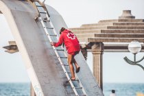 Rückansicht eines jungen Lebensretters im roten Anzug, der die Treppe am Strand hinuntersteigt — Stockfoto