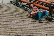 Low angle view of smiling man in casual clothes leaning on street steps and holding smartphone in hand — Stock Photo