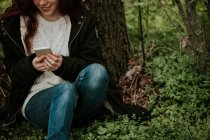 Cortar menina gengibre sentado à árvore e navegação smartphone — Fotografia de Stock
