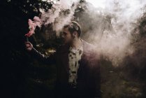 Portrait de jeune homme posant avec une bougie de fumée rose dans les bois — Photo de stock