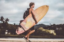Vista lateral de un joven surfista con mochila recorriendo la playa con una tabla de surf - foto de stock