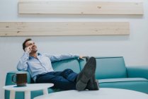 Veduta di un giovane uomo d'affari seduto sul divano, che parla al cellulare, alza lo sguardo e ride. Copyspace — Foto stock