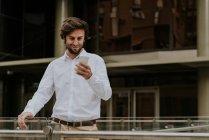 Retrato de homem de negócios sorridente em camisa branca olhando para smartphone na mão em cena urbana — Fotografia de Stock