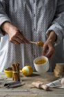 Sección media de la hembra sosteniendo la cuchara de miel con taza en las manos sobre la mesa rústica de la cocina con ingredientes y utensilios de cocina - foto de stock