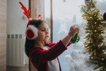 Vista laterale di adorabile ragazza che indossa cuffie di pelliccia di Natale con corna che mettono bagattelle sull'albero di Natale decorativo — Foto stock