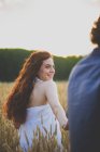 Back view of girl with curvy red hair walking on rye field and looking over shoulder at boyfriend — Stock Photo
