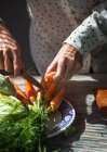 Vista de cerca de las manos femeninas de corte con cuchillo zanahorias hojas sobre plato en la mesa de la cocina - foto de stock