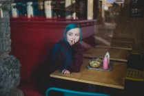 Ritratto di ragazza con i capelli blu seduta a tavola con yogurt e ciotola di cereali su vassoio e guardando la finestra — Foto stock
