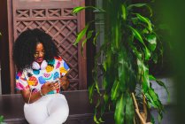 Портрет девушки в красочной футболке, сидящей рядом с цветочным горшком и болтающей на смартфоне — стоковое фото