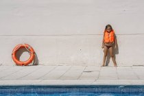 Chica rubia con chaleco salvavidas naranja de pie en la piscina y mirando a la cámara . - foto de stock