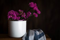 Cravos roxos frescos na caneca no fundo escuro com toalha de cozinha — Fotografia de Stock