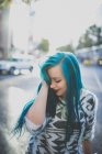Portrait of young girl wearing soft sweater smoothing her blue straight hair and looking down at street scene — Stock Photo