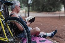 Вид сбоку пожилого человека, читающего книгу, сидя на земле в лесу рядом с велосипедом — стоковое фото