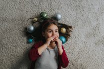Above view of girl laying on carpet floor with christmas baubles around head — Stock Photo