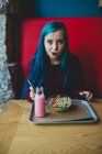 Портрет синеволосой девочки-подростка, сидящей за столиком кафе с йогуртом и миской красочных злаков на подносе и смотрящей в камеру — стоковое фото