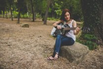 Ritratto di sorridente ragazza lentigginosa seduta sulla pietra al bosco e che suona la chitarra — Foto stock
