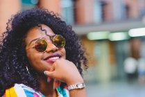 Ritratto di donna in occhiali da sole specchiati con acconciatura afro sorridente alla macchina fotografica con mano sul mento — Foto stock