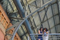 Vista a basso angolo di due donne appoggiate sulla ringhiera del balcone e utilizzando il telefono cellulare sotto la costruzione del tetto in ferro . — Foto stock