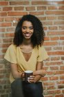 Smiling brunette woman with cup looking at camera on the background of brick wall. Vertical indoors shot — Stock Photo
