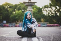 Portrait of blue haired girl sitting on asphalt road and flirtatious looking at camera — Stock Photo