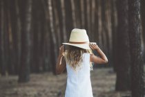 Rear  view of little girl wearing hat and white dress posing on background of forest. — Stock Photo