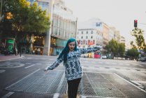 Retrato de chica de pelo azul caminando con los brazos medio levantados y mirando hacia abajo a la escena urbana - foto de stock
