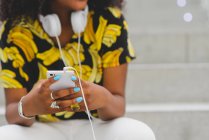 Mittelteil einer Afro-Frau mit Kopfhörern um den Hals, die auf einer Treppe sitzt und ihr Smartphone in der Hand hält — Stockfoto