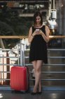 Retrato de mujer morena con maleta roja apoyada en parapeto y usando smartphone - foto de stock