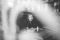 Retrato de menina sorridente com sentado na mesa de café com smartphone e olhando para a câmera — Fotografia de Stock