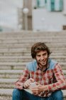 Retrato de homem sorridente em camisa quadriculada sentado em degraus de rua e usando smartphone — Fotografia de Stock