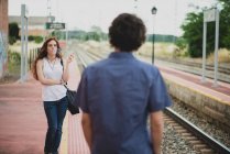 Вид сзади на мужчину, смотрящего на курящую девушку с вьющимися рыжими волосами на железнодорожной платформе — стоковое фото