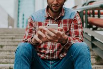 Mann im karierten Hemd surft Smartphone, während er auf Stufen sitzt — Stockfoto