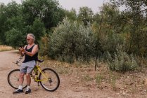 Retrato del hombre mayor apoyado en la bicicleta y el teléfono inteligente de navegación en el parque - foto de stock