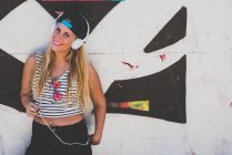 Portrait of attractive young blonde woman listening to music in headphones against graffiti wall. — Stock Photo