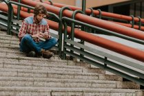 Portrait of smiling bearded man in casual clothes sitting on street steps and using phone — Stock Photo