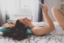 Chica acostada en la cama en la espalda con teléfono inteligente en las manos - foto de stock