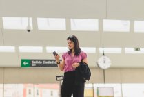 Retrato de mujer de pie en el vestíbulo de la estación con equipaje y mirando el teléfono inteligente en la mano - foto de stock