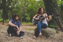 Ritratto di uomo seduto a terra che ascolta una ragazza seduta sul rock e suona la chitarra nel bosco — Foto stock