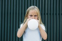 Portrait of serious little girl looking at camera while blowing white balloon at street. — Stock Photo