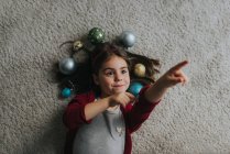 Above view of girl laying on carpet floor with christmas baubles around head and pointing with finger — Stock Photo