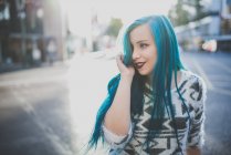 Portrait of young girl wearing soft sweater smoothing her blue straight hair and looking aside at street scene — Stock Photo