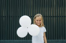 Chica sonriendo a la cámara mientras sostiene tres globos blancos - foto de stock
