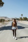 Tiro de longitud completa de la joven mujer con estilo que camina por la calle a la luz del sol en el fondo de colinas rocosas. - foto de stock