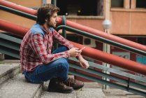 Vista lateral do homem barbudo em roupas casuais sentado em degraus de rua com smartphone e olhando para longe — Fotografia de Stock