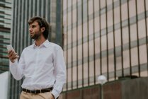 Retrato de empresário confiante em camisa branca segurando smartphone e olhando para a cena urbana — Fotografia de Stock