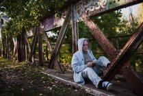 Viejo con capucha sentado en la construcción de un puente de metal y mirando algo en sus manos. Espacio de copia - foto de stock