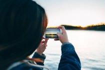 Vue sur l'épaule de la femme prenant une photo du coucher du soleil sur smartphone . — Photo de stock